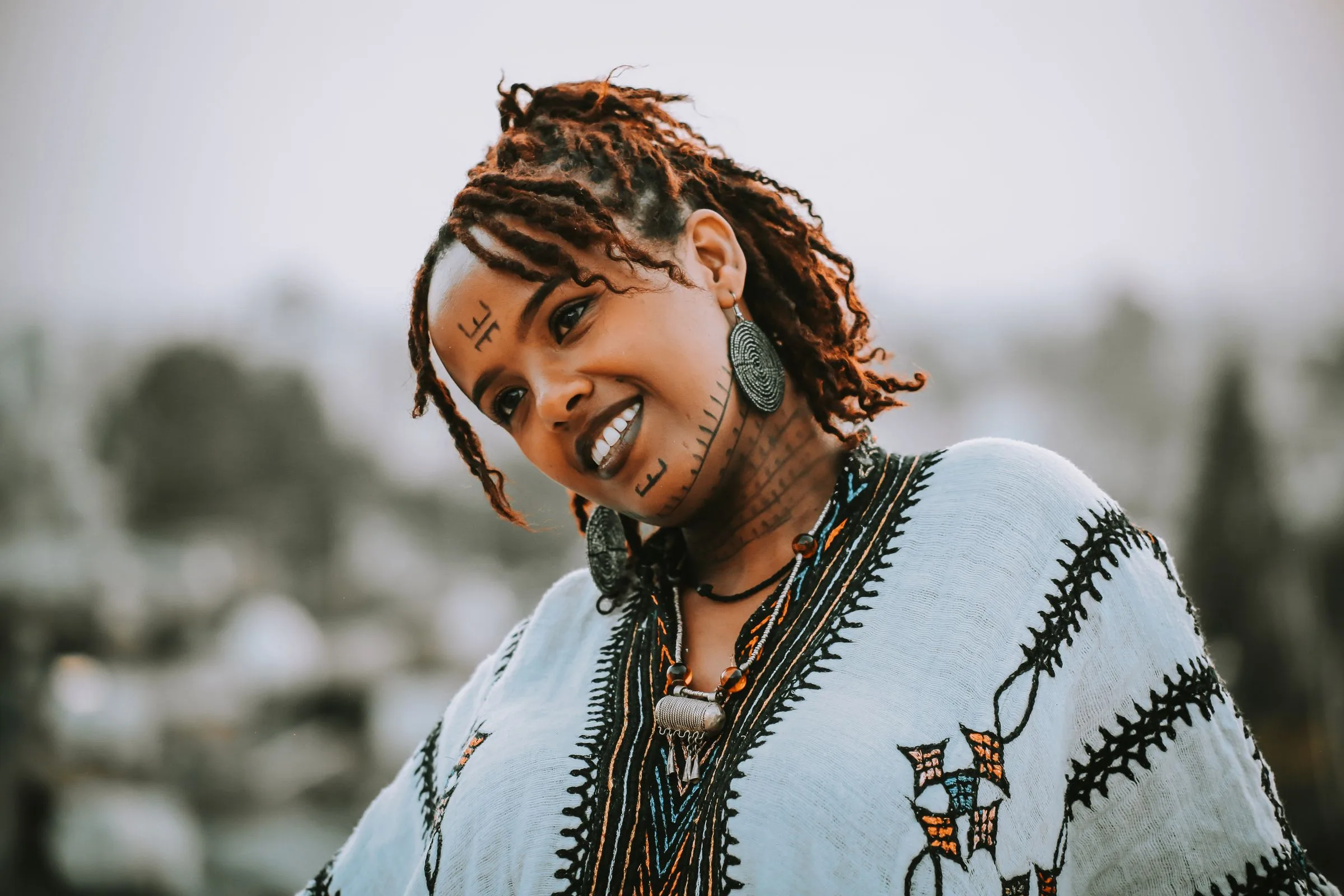 A smiling Ethiopian woman in an embroidered white habesha kemis, with traditional facial tattoos