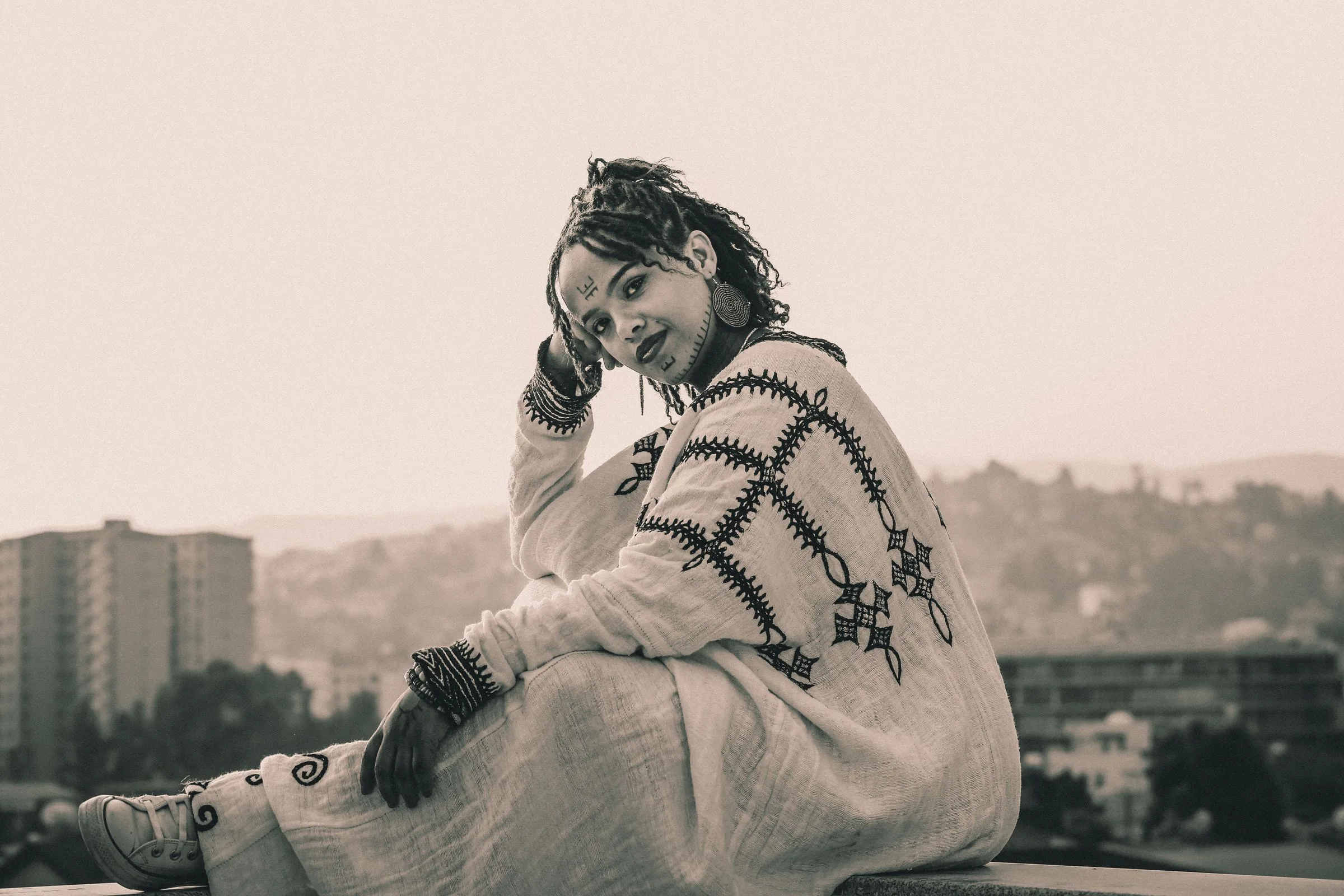 A young Ethiopian woman in a white dress embroidered with black crosses sits on a rooftop in Addis Ababa, with the city behind her