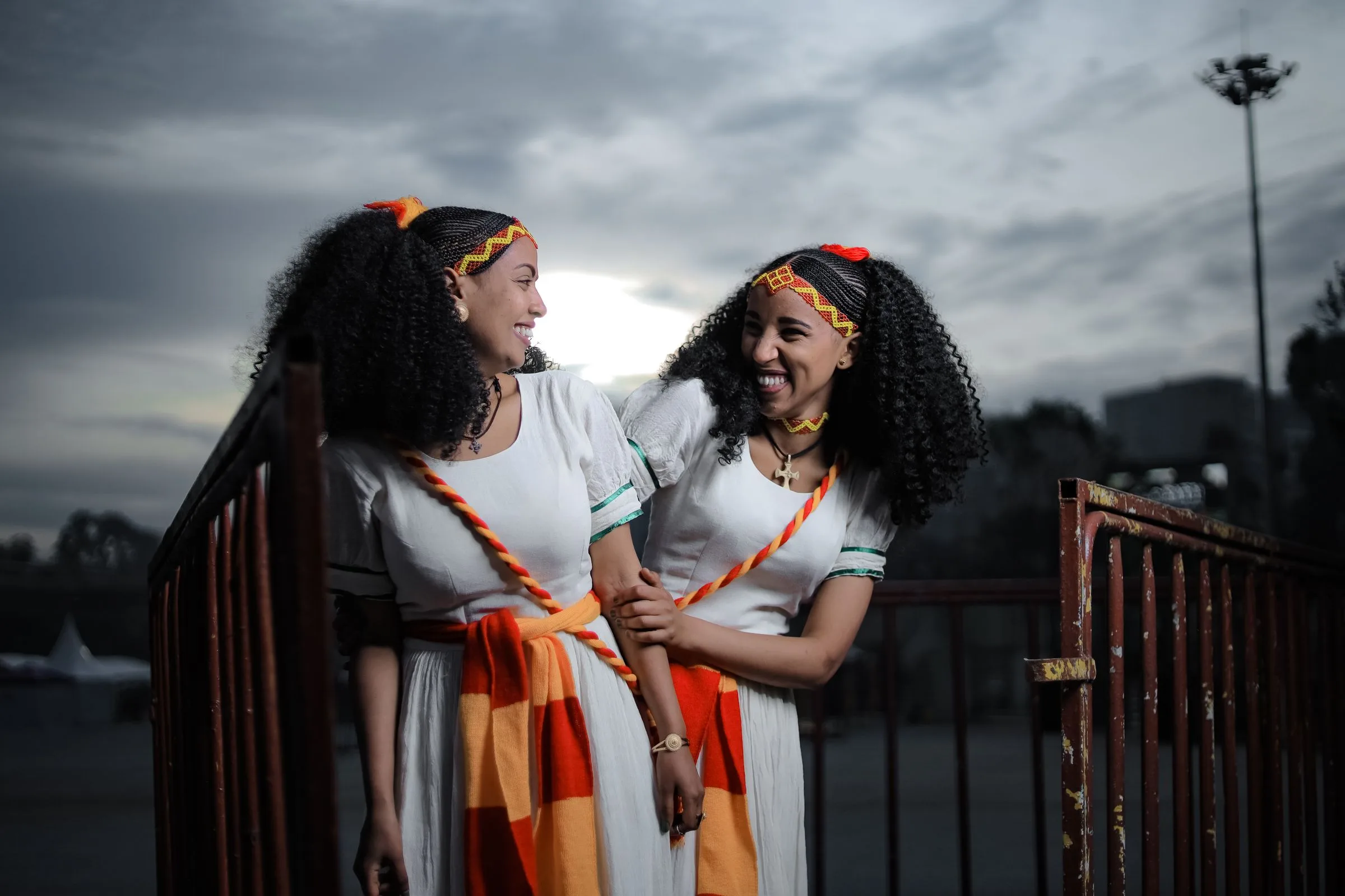Two young Ethiopian women in white habesha dresses with orange sashes laugh together by a railing in Addis Ababa
